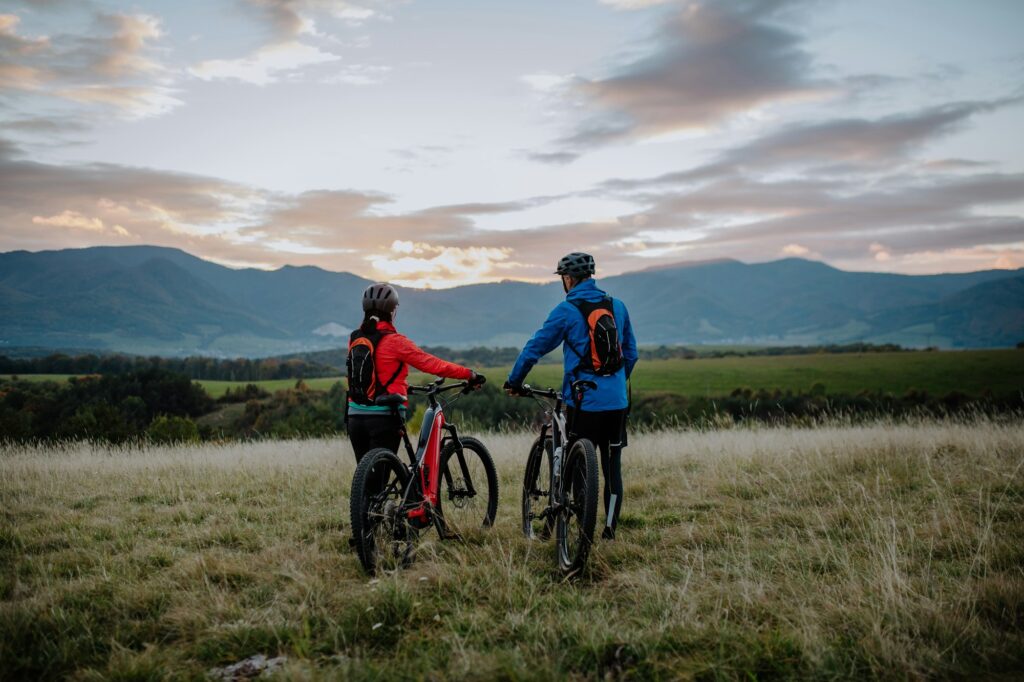 Rear view of senior couple bikers walking and pushing e-bikes outdoors in forest in autumn day